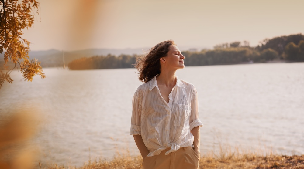 Beautiful young relaxed woman in white blouse enjoying nature breathing fresh air meditating on the river on an autumn day