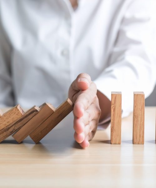 A person in white shirt stops falling wooden dominoes with their hand, symbolizing intervention, risk management, and prevention in business or personal context