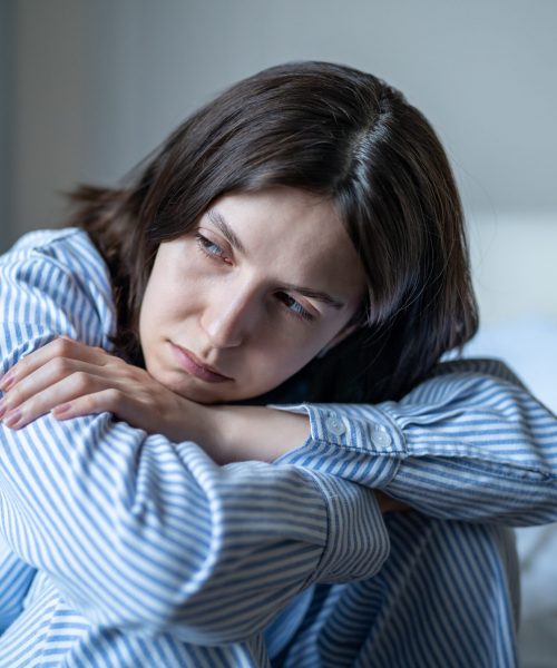 Tired female struggling with depression, solitude, feeling hopeless, suffering from sadness, exhaustion, psychological problem. Upset female sits on bed in nightwear overwhelmed deep negative thoughts