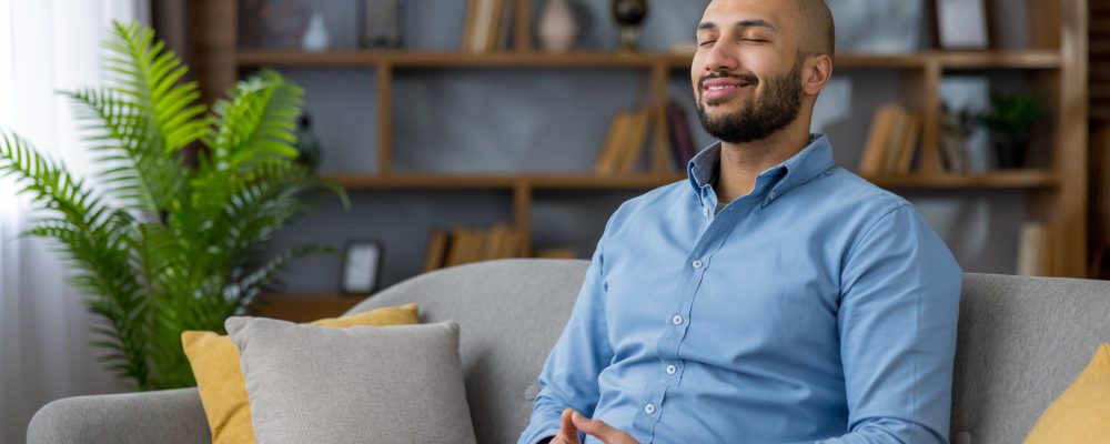 Young man sitting on a grey couch with eyes closed and a gentle smile, enjoying a calm, mindful moment of relaxation and inner peace at home