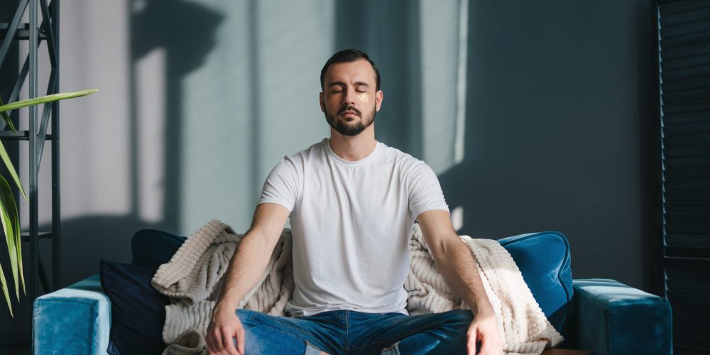 Focused man meditating in lotus pose breathing deep and slowly wearing casual clothes sitting on comfortable coach relaxing at home. Feeling free from stress.