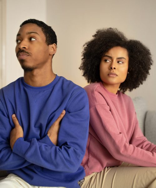 Young African American couple faces marriage crisis, husband and wife sitting in sulking silence after conflict, turning back with discontented faces on sofa at home interior