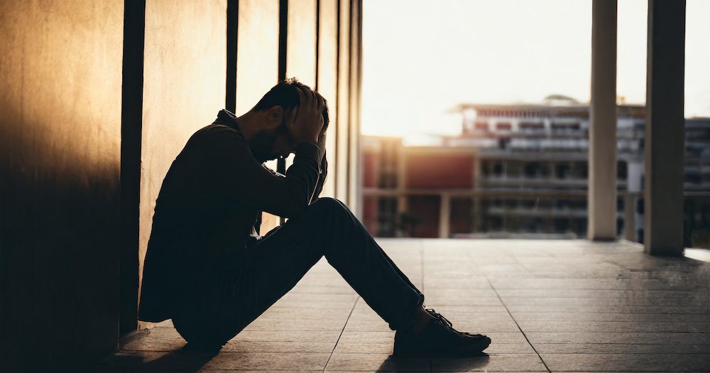 Silhouette of despondent man, head in hands, seated on residence walkway