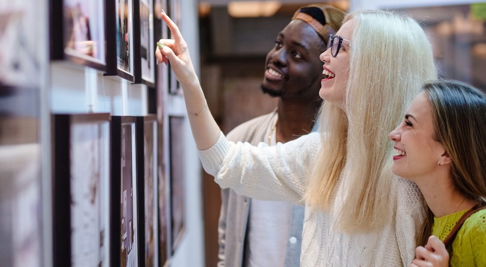 Three young people hipster students with different skins looking happy together, talkng, looking at picture on wall, spending free time together in art photo gallery. Multi ethnic, friendship concept.